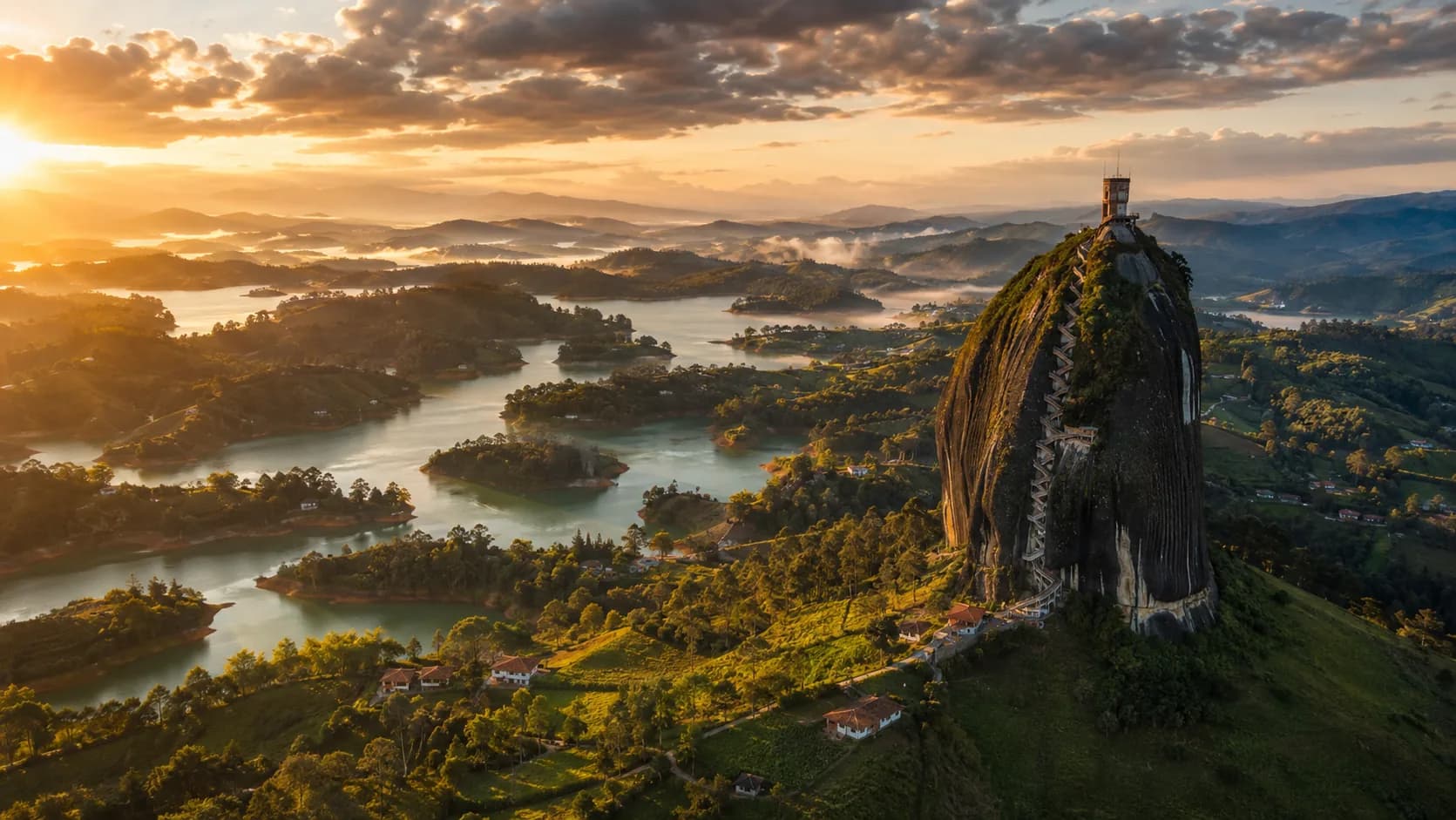 Vista aérea de la Piedra del Peñol al atardecer, con su escalera en zigzag subiendo por la fisura, sobre el embalse de Guatapé y las colinas verdes del Oriente antioqueño.