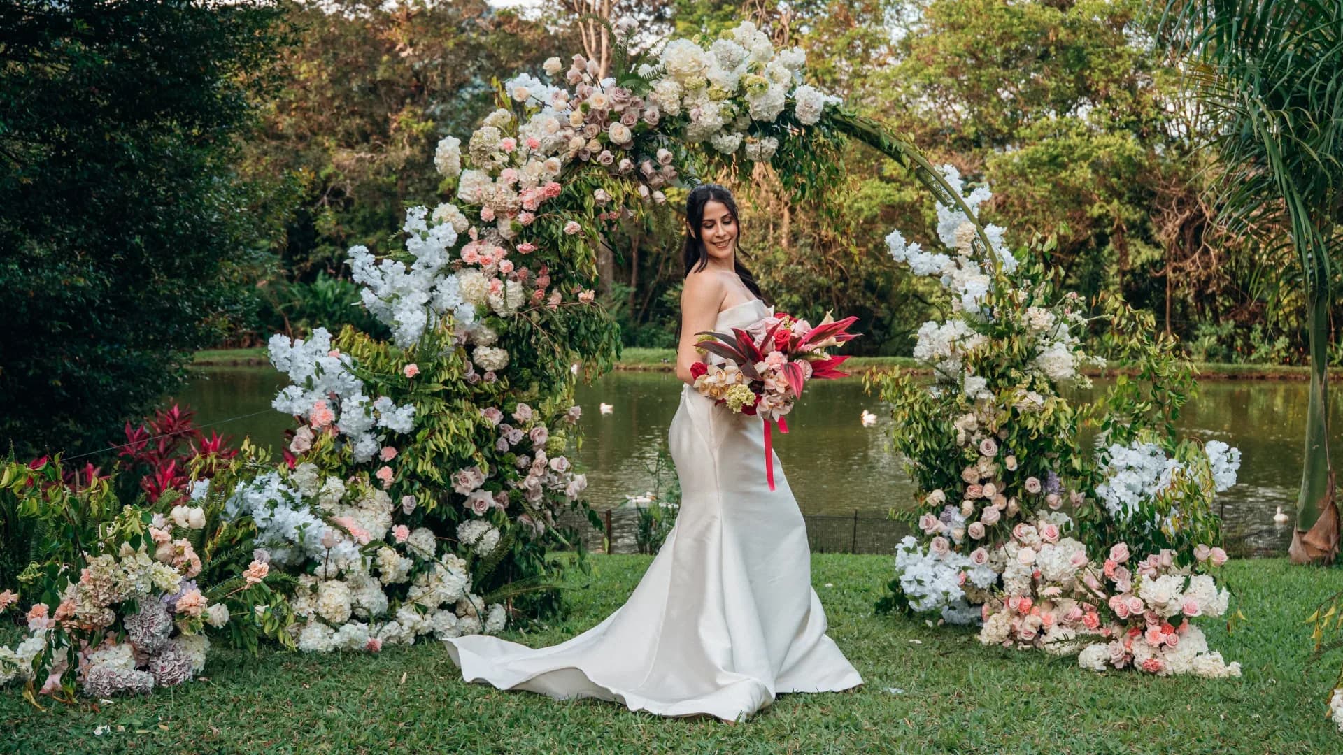 Una novia con su ramo bajo un arco de flores blancas y rosadas, junto a un lago en Girardota, Antioquia.