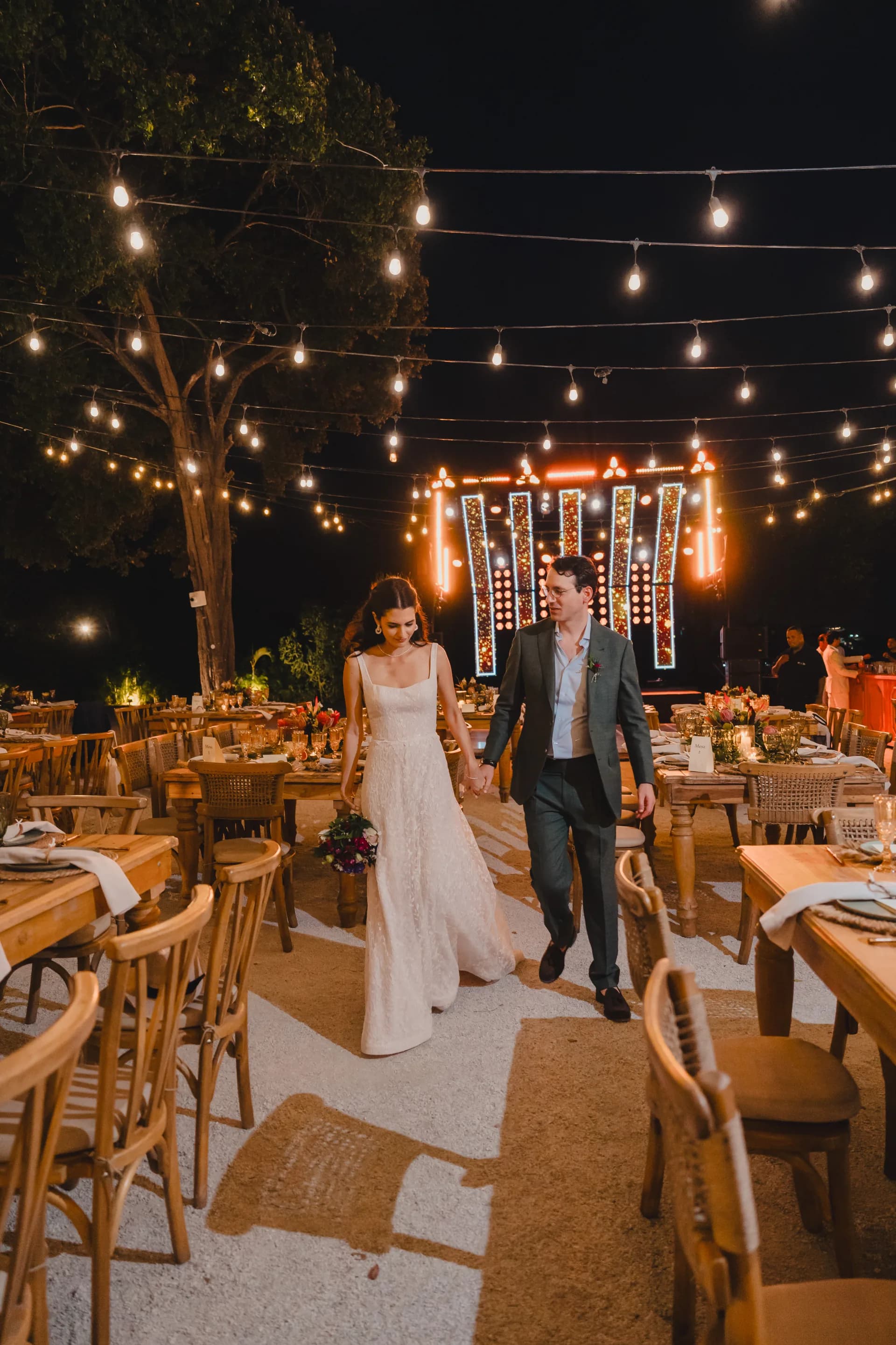 Los novios caminan de la mano por la recepción de noche, bajo guirnaldas de luces